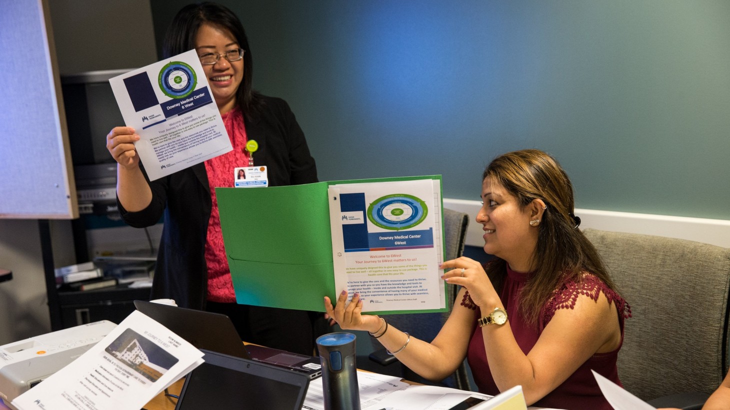 two women at a meeting table, holding up documents 